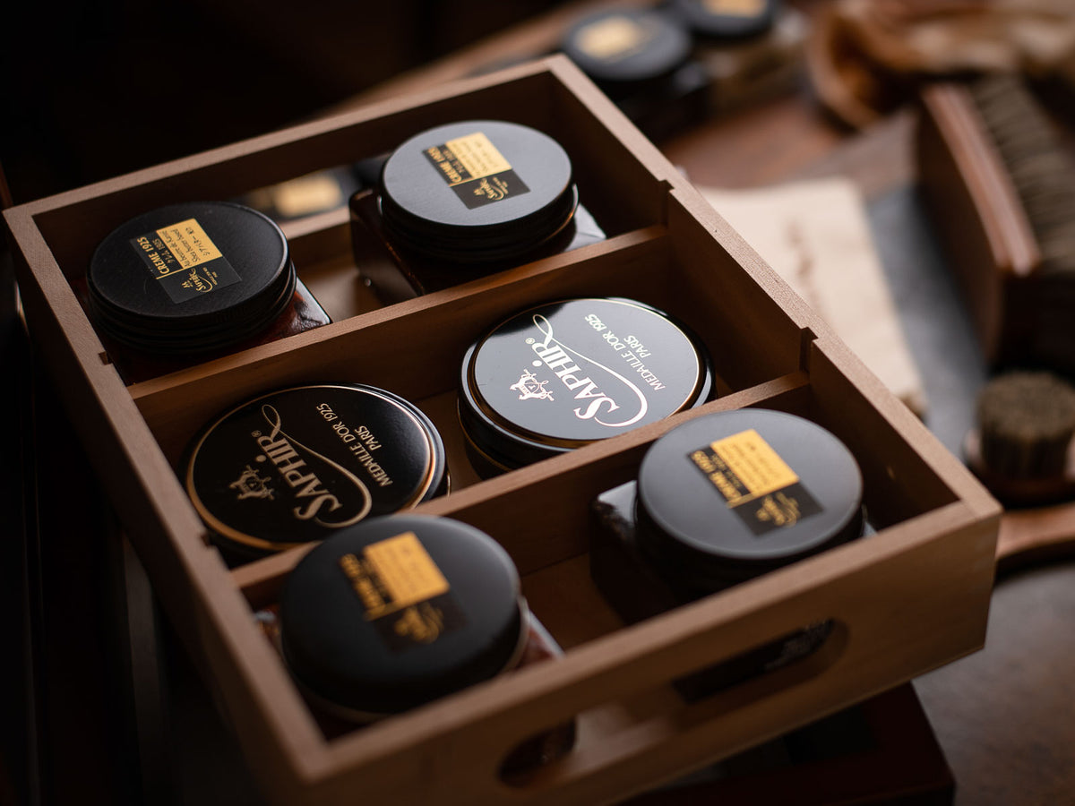 Close-up of shoe polish tins in a wooden organizer. Dark polish tins with gold labels read "Saphir" and "Crème 1925." Moody lighting.
