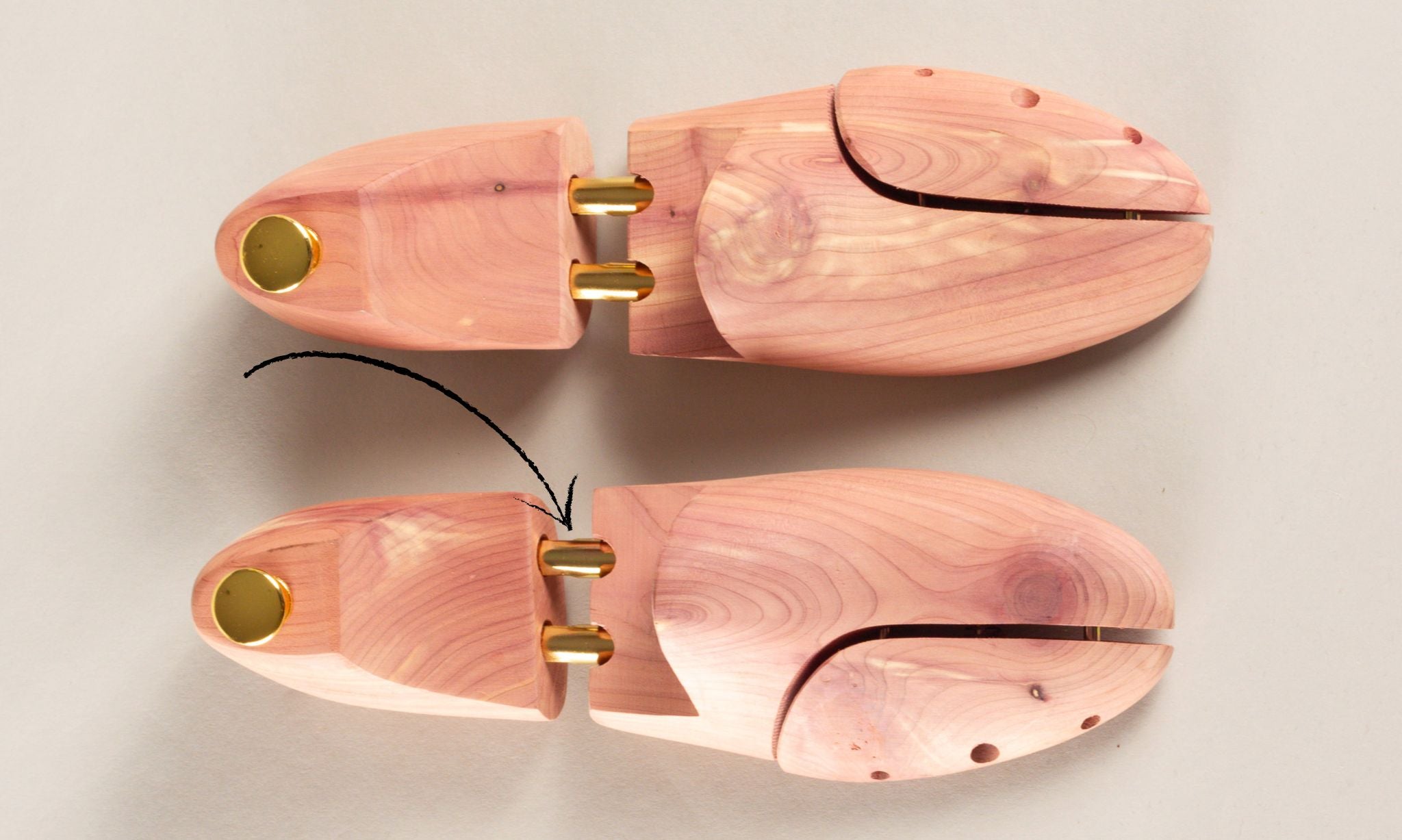 Two cedar shoe trees lying on a light gray surface, showing their two-part construction. A black curved arrow points towards the metal hinge.