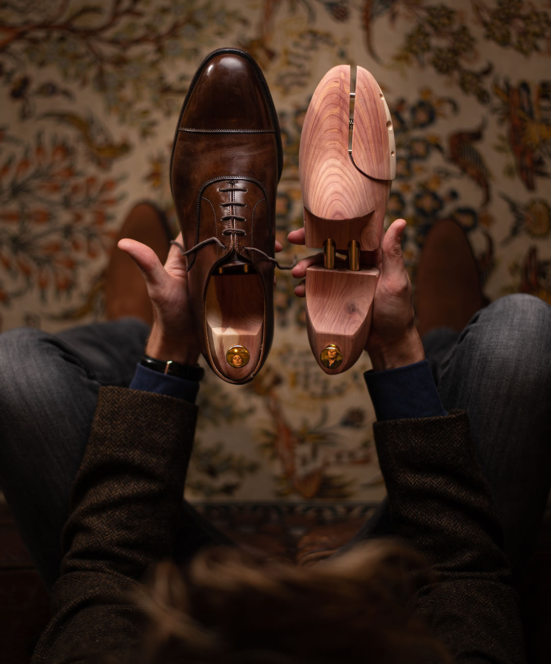 A person holding a brown leather dress shoe in one hand and a wooden shoe tree in the other, sitting on a patterned carpet with legs crossed, seen from above.